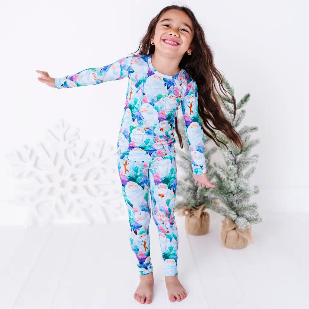 A smiling child with one arm stretched out, barefoot in a long sleeve pajama set with a print of friendly white yetis among teal, blue, and purple pine trees, beside small frosted trees.