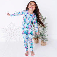 A smiling child with one arm stretched out, barefoot in a long sleeve pajama set with a print of friendly white yetis among teal, blue, and purple pine trees, beside small frosted trees.