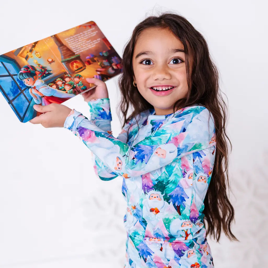 A child with a wide smile holding an open storybook up high, in a long sleeve pajama top with a print of friendly white yetis among teal, blue, and purple pine trees.