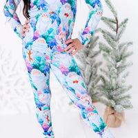 Close up of a child with hands on hips, wearing a long sleeve pajama top and matching pants in a print of friendly white yetis among teal, blue, and purple pine trees.