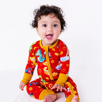 A baby with a big open mouth smile sitting on a white floor in a zip up pajama with mustard yellow trim and cuffs in a red print of zodiac animals and gold clouds.