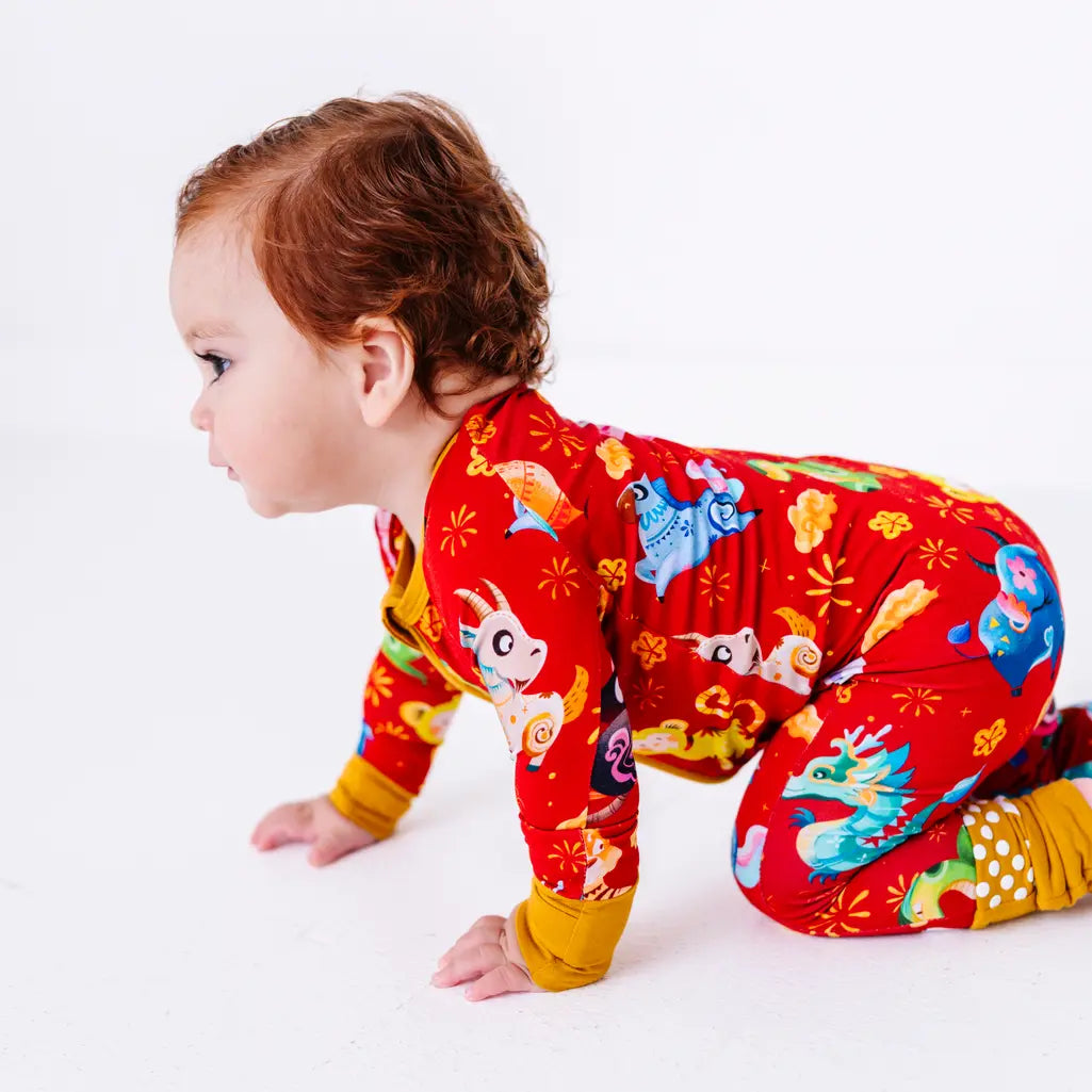 Side view of a baby crawling on a white floor in a zip up pajama with mustard yellow cuffs and grip dots on the feet, in a red print of zodiac animals and gold clouds.