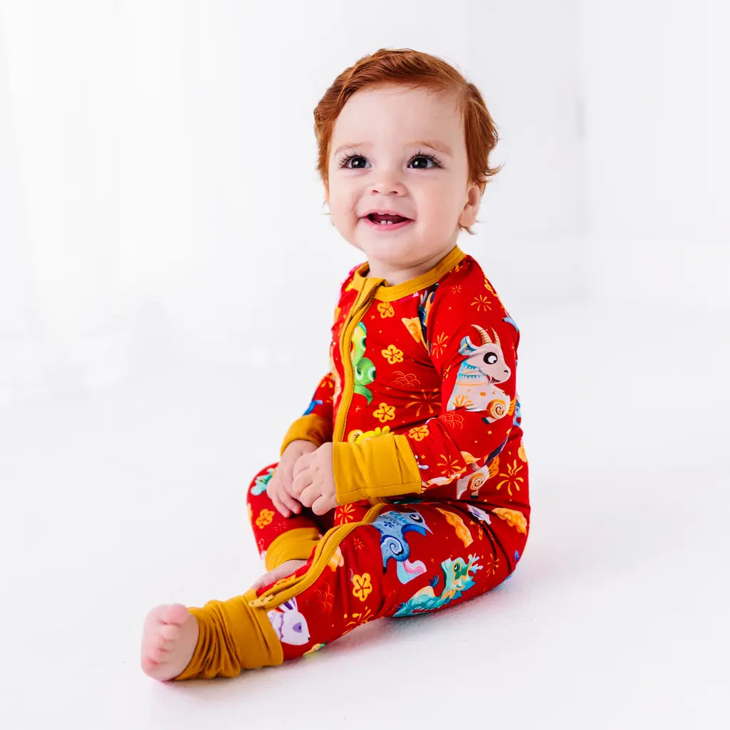 A baby smiling at the camera while sitting with one leg stretched out, showing the leg zipper and folded foot cuff of a pajama in a red print of zodiac animals and gold clouds.