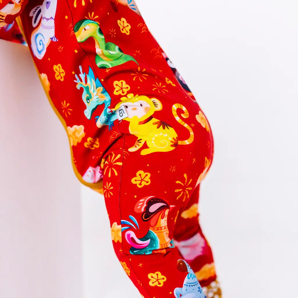 Close up of the leg and hip of a zip up baby pajama with mustard yellow trim along the seam, in a red print of zodiac animals and gold clouds.