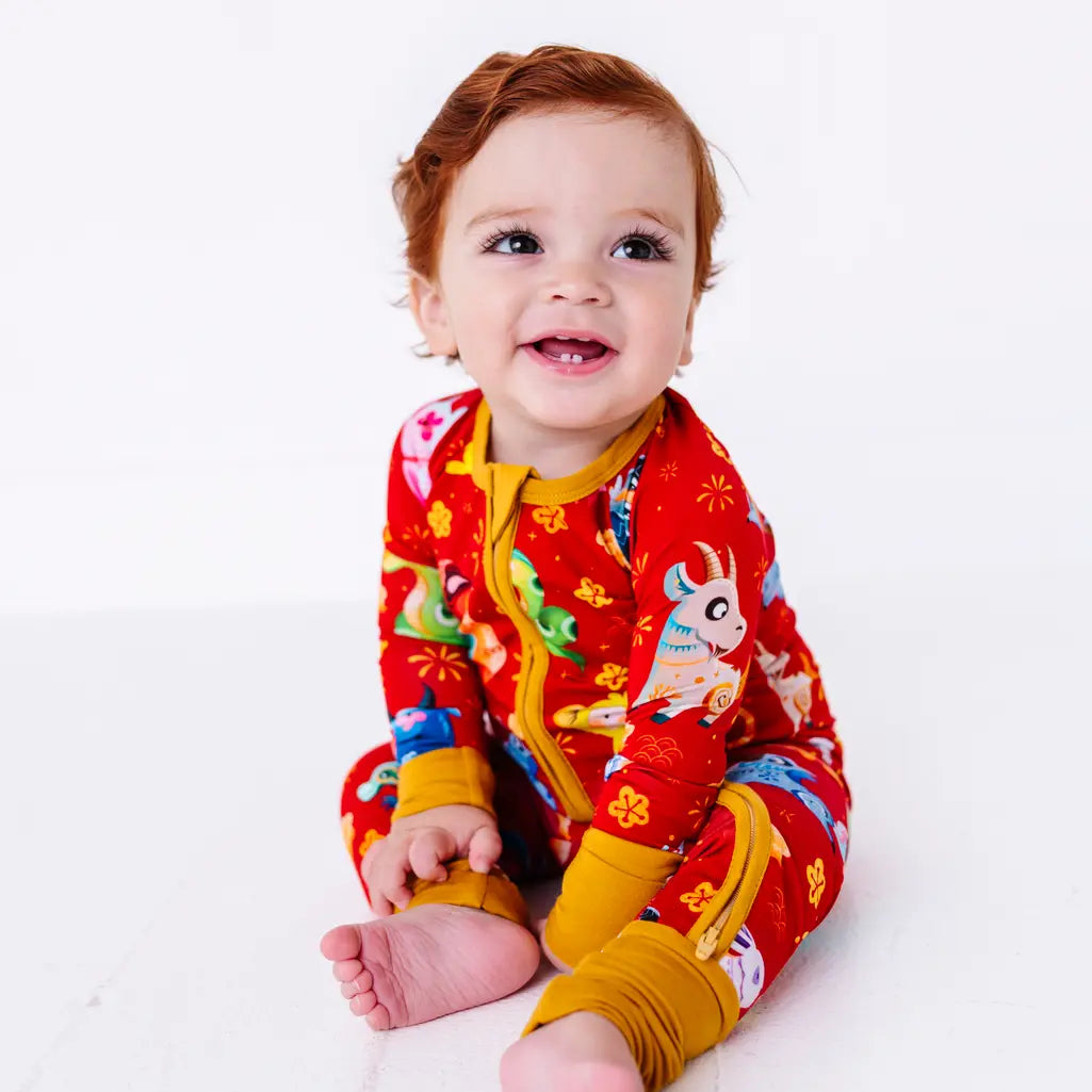 A baby sitting barefoot on a white floor and smiling at the camera in a zip up pajama with mustard yellow trim and cuffs in a red print of zodiac animals and gold clouds.