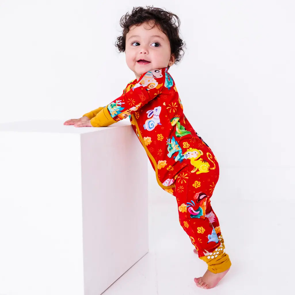 A smiling baby standing barefoot and leaning on a white block while looking back at the camera, in a zip up pajama in a red print of zodiac animals and gold clouds.