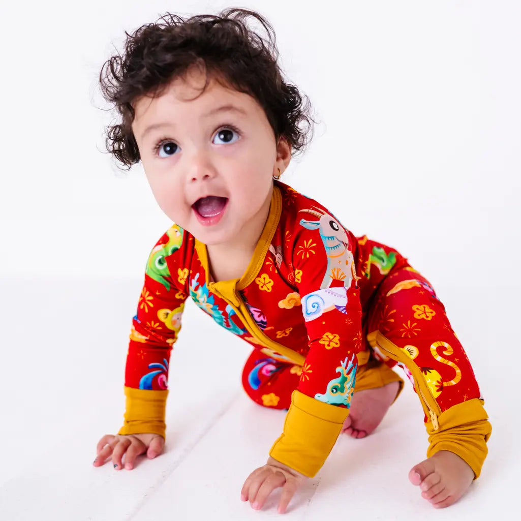 A baby crawling toward the camera with an open mouth smile in a zip up pajama with mustard yellow cuffs in a red print of zodiac animals and gold clouds.