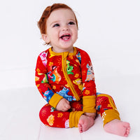 A baby laughing and looking up while sitting on a white floor, in a zip up pajama with mustard yellow trim and cuffs in a red print of zodiac animals and gold clouds.