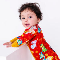 A baby smiling at the camera while leaning on a white block, in a zip up pajama with mustard yellow cuffs in a red print of zodiac animals and gold clouds.