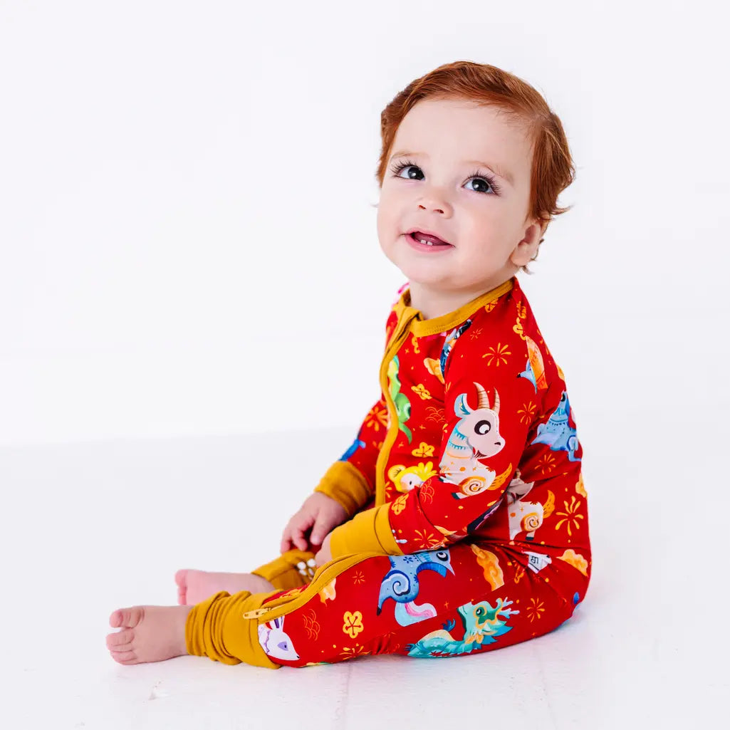 Side view of a baby sitting and looking up with a small smile, in a zip up pajama with mustard yellow trim in a red print of zodiac animals and gold clouds.