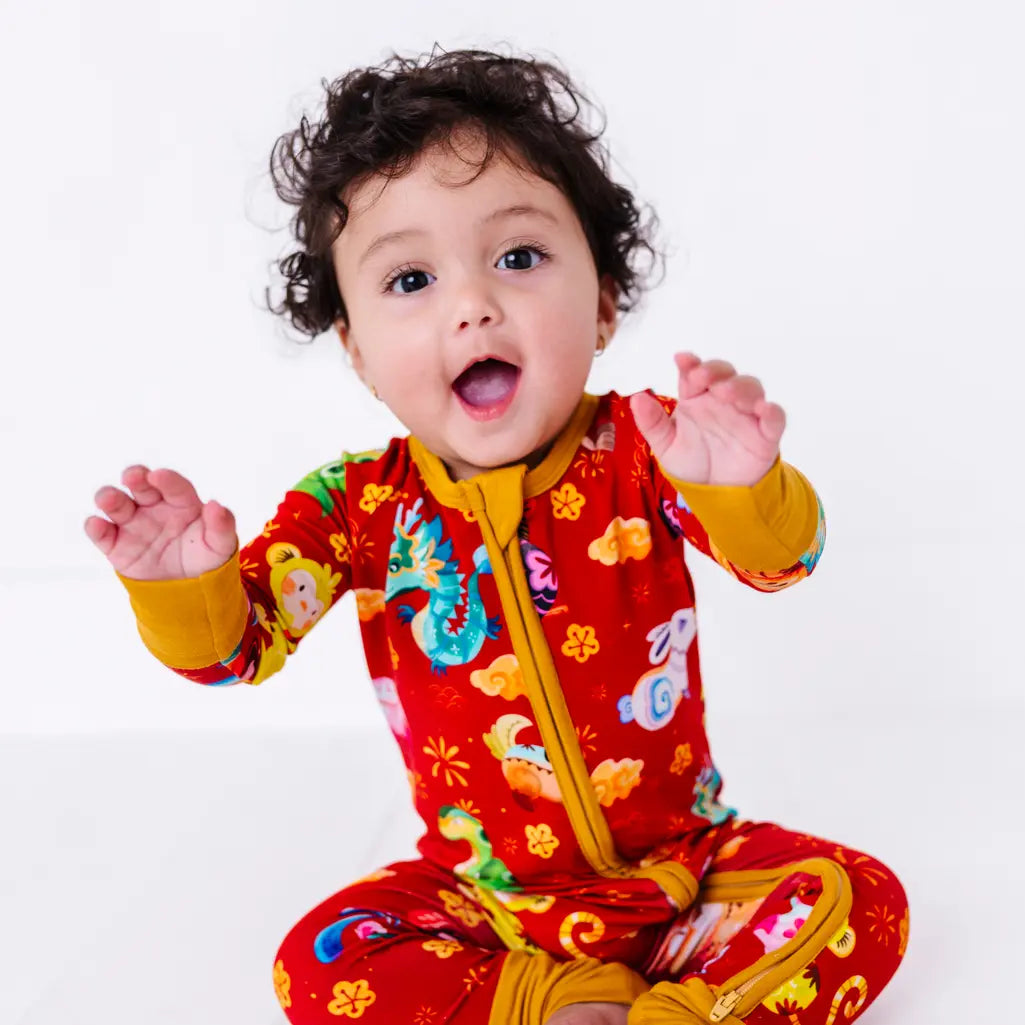 A baby with both hands raised and an open mouth smile, sitting in a zip up pajama with mustard yellow cuffs in a red print of zodiac animals and gold clouds.