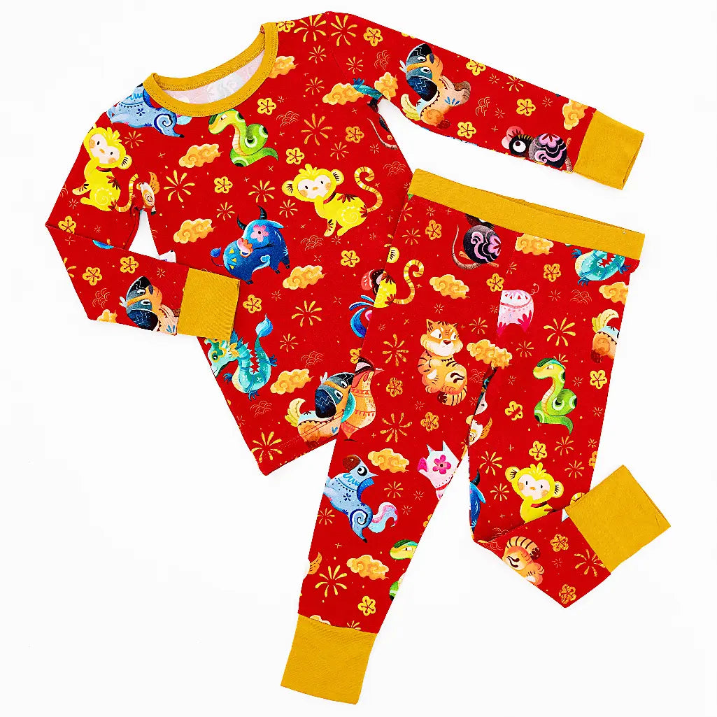 Flat lay of a long sleeve pajama top and matching pants with mustard yellow cuffs in a red print of zodiac animals and gold clouds on a white background.