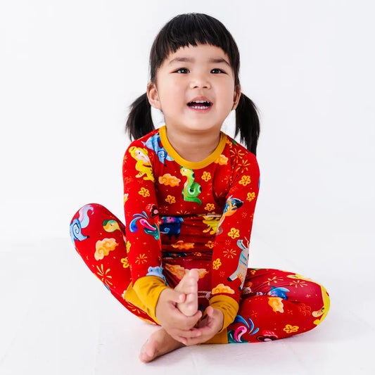 A child with a big smile sitting cross legged and holding one bare foot, in a long sleeve pajama top and matching pants with mustard yellow cuffs in a red print of zodiac animals and gold clouds.
