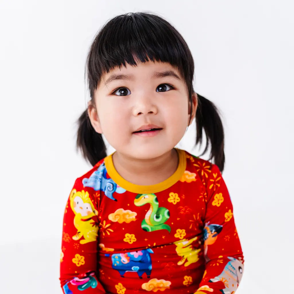 Close up portrait of a child looking up with a soft smile, in a long sleeve pajama top with a mustard yellow collar in a red print of zodiac animals and gold clouds.