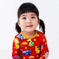 Close up portrait of a child looking up with a soft smile, in a long sleeve pajama top with a mustard yellow collar in a red print of zodiac animals and gold clouds.
