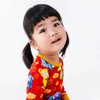 A child with head tilted looking up with a smile, shown from the chest up, in a long sleeve pajama top with a mustard yellow collar in a red print of zodiac animals and gold clouds.