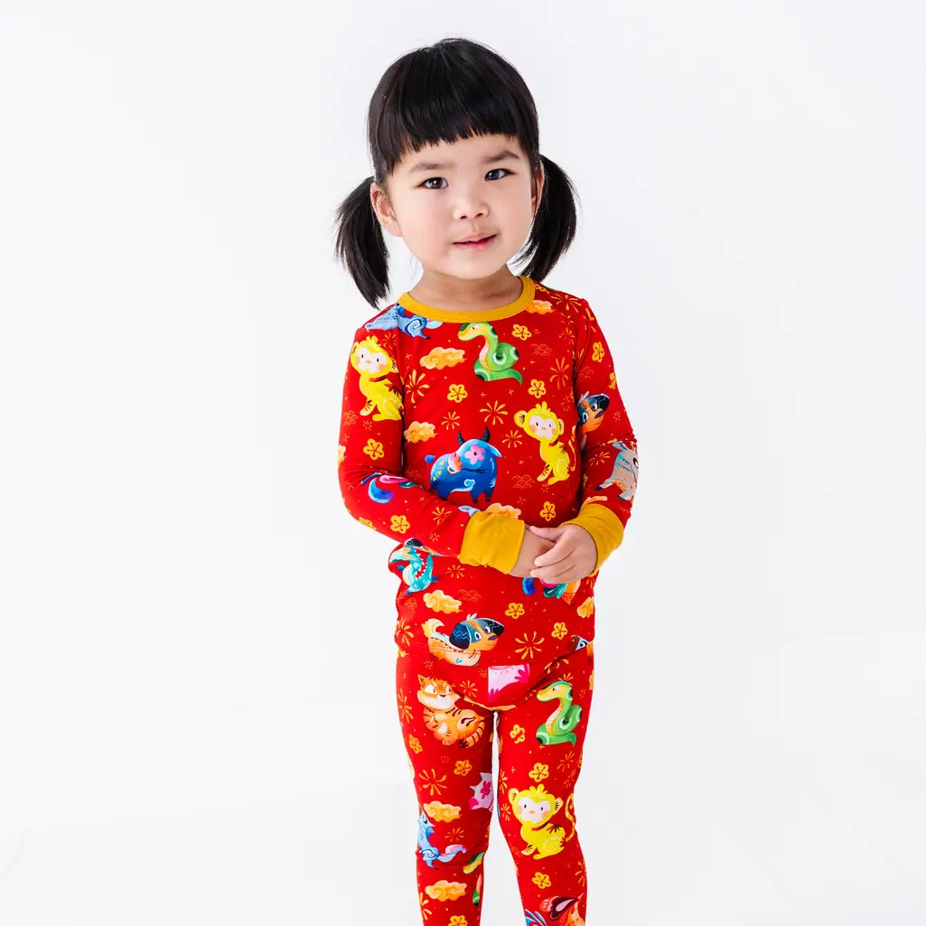 A child standing with hands clasped in a long sleeve pajama top and matching pants with mustard yellow cuffs in a red print of zodiac animals and gold clouds.