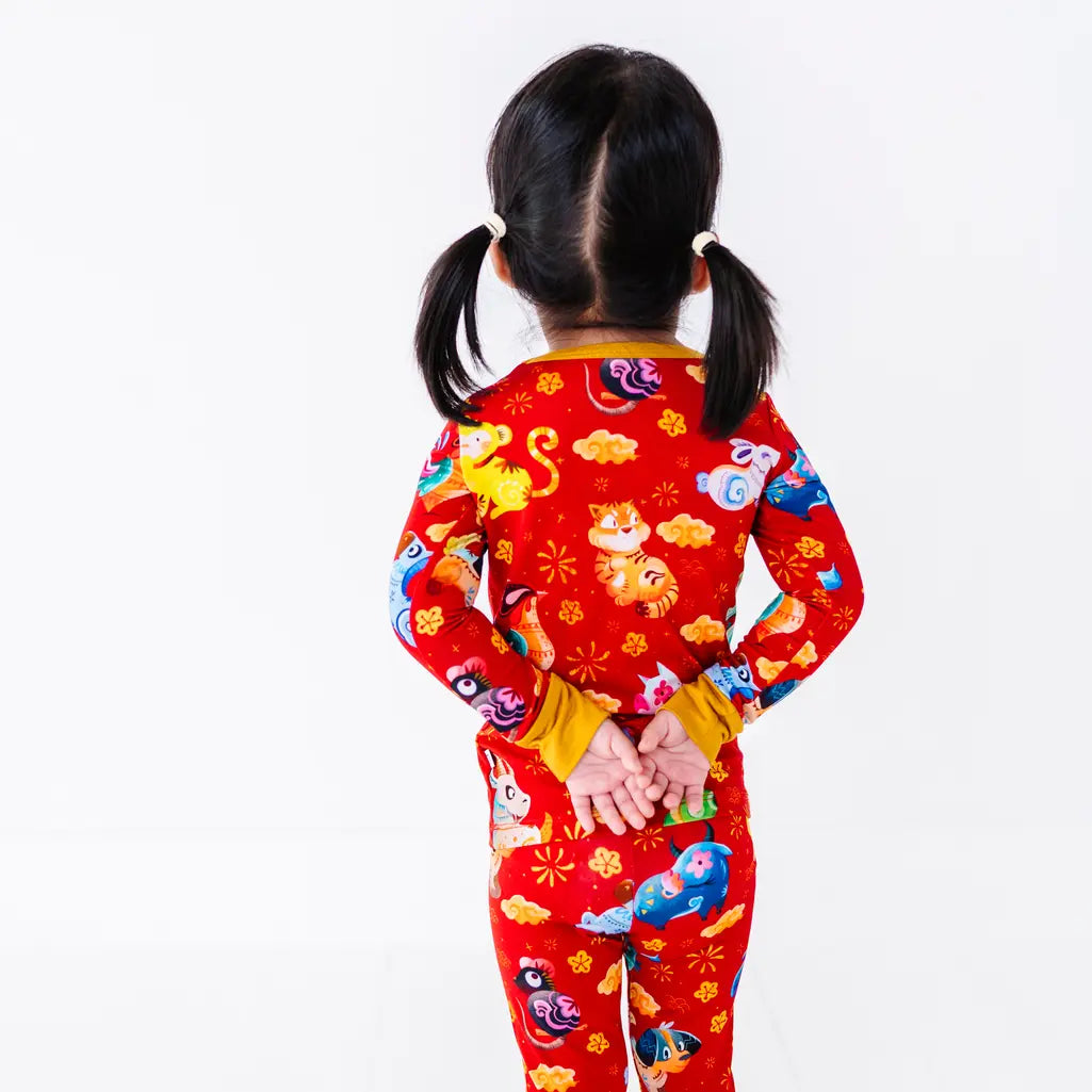 Back view of a child standing with hands behind the back, in a long sleeve pajama top and matching pants in a red print of zodiac animals and gold clouds.