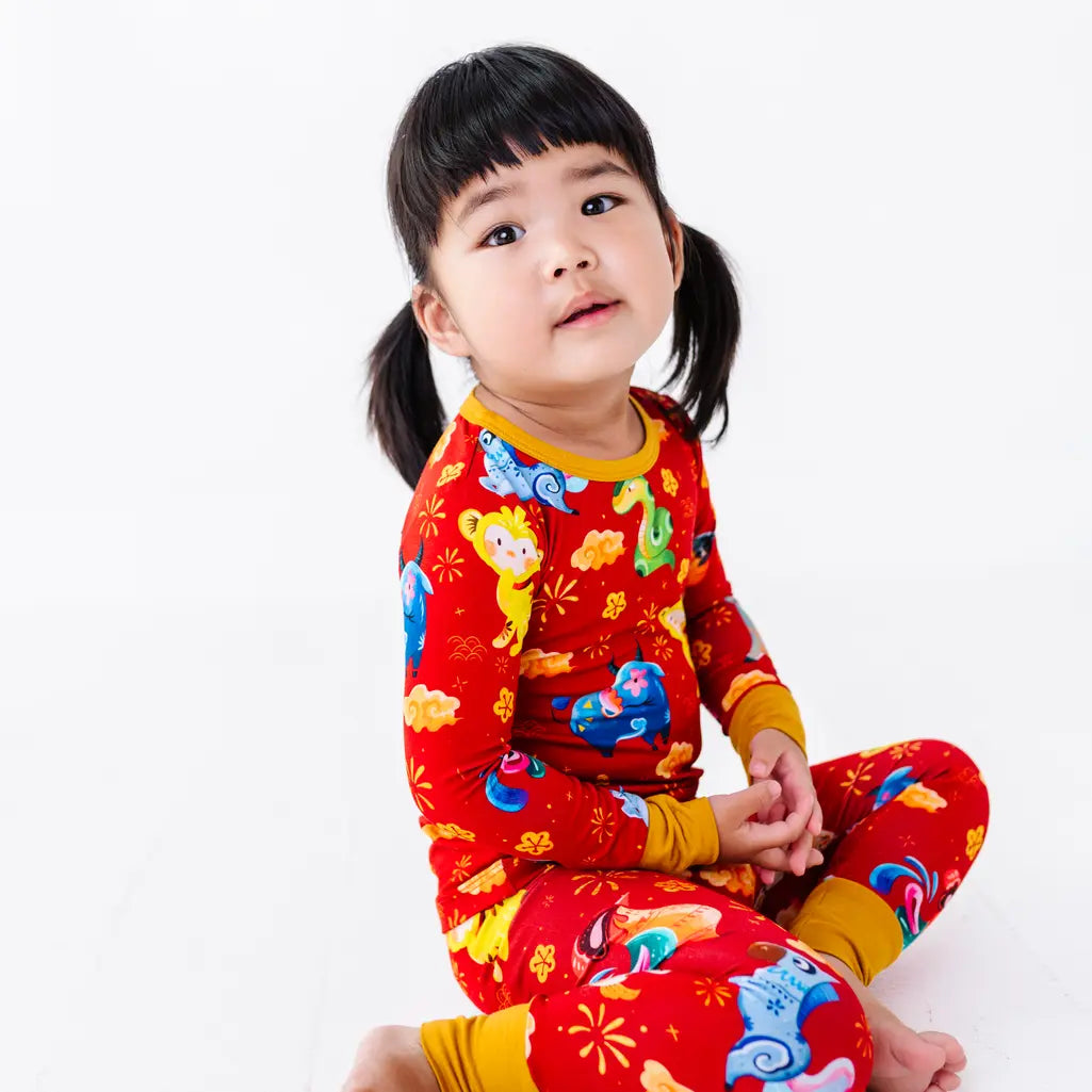 A child sitting barefoot with hands clasped and looking up at the camera, in a long sleeve pajama top and matching pants in a red print of zodiac animals and gold clouds.