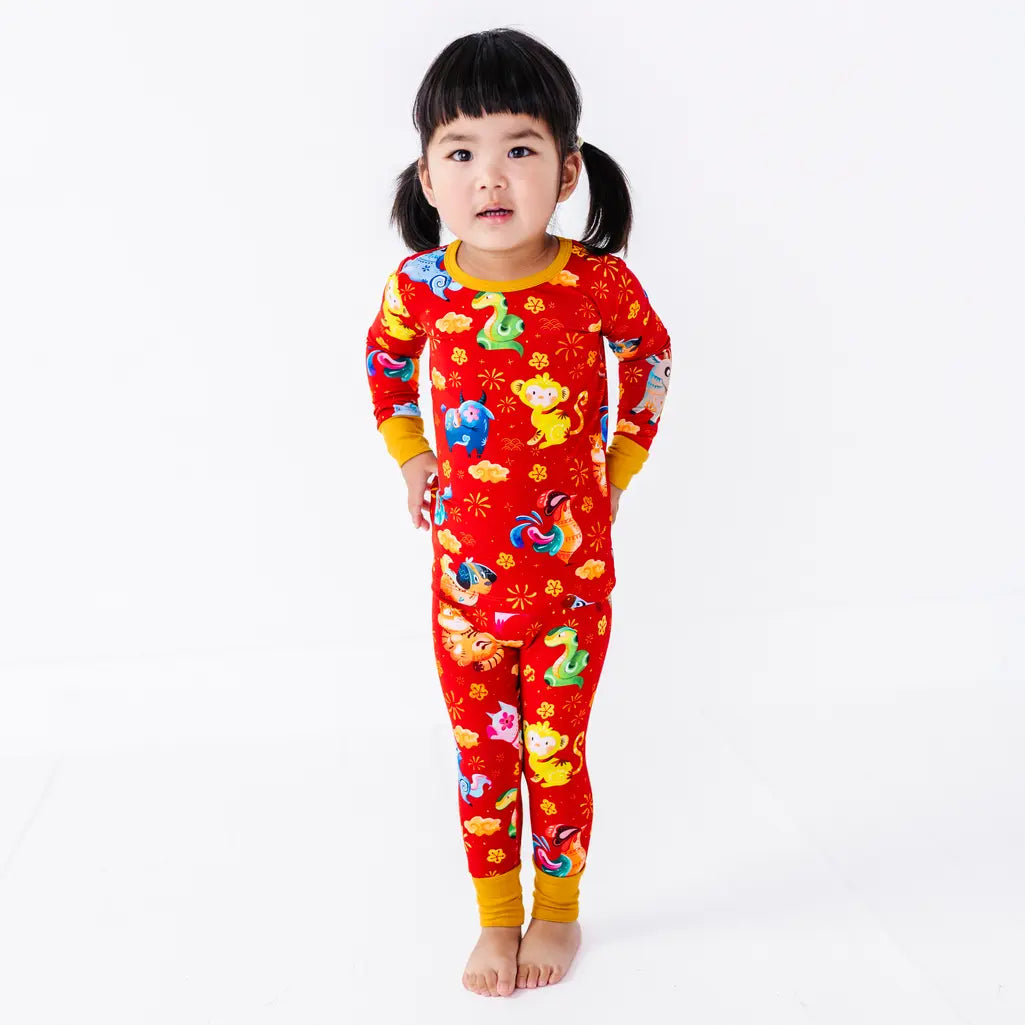 A child standing barefoot with hands by the hips, shown full length, in a long sleeve pajama top and matching pants in a red print of zodiac animals and gold clouds.