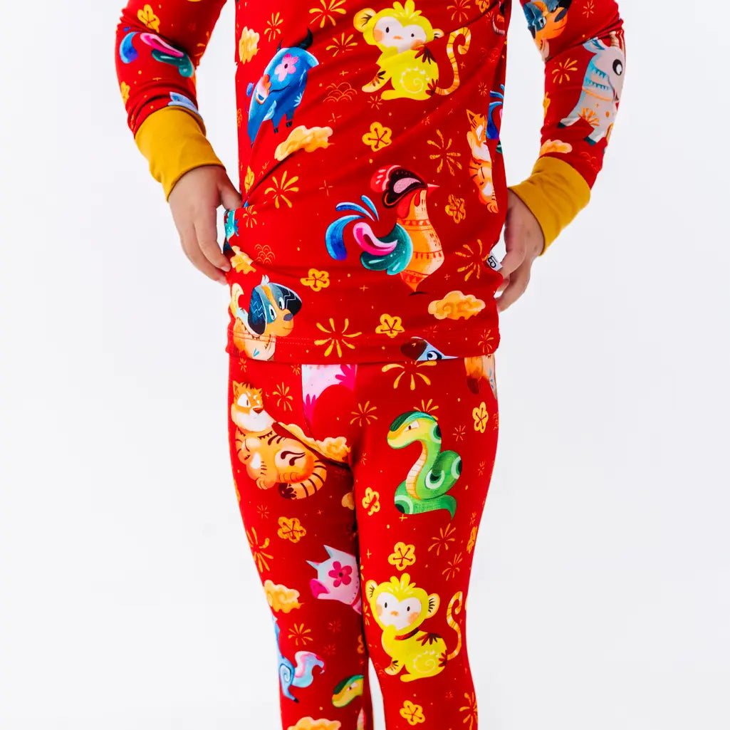 Close up of a child with hands on hips, showing the hem of a long sleeve pajama top and matching pants in a red print of zodiac animals and gold clouds.