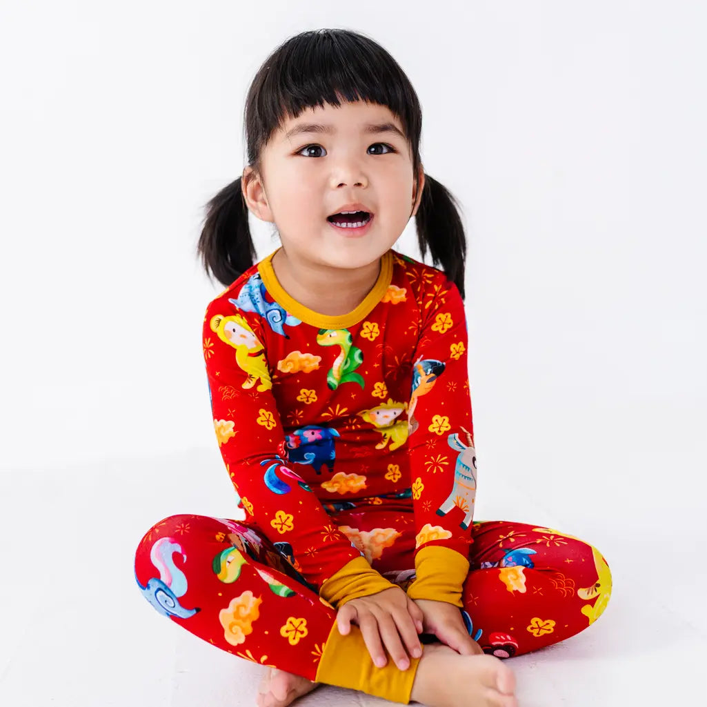 A child sitting cross legged and looking at the camera with an open mouth, in a long sleeve pajama top and matching pants in a red print of zodiac animals and gold clouds.