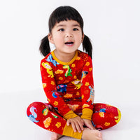 A child sitting cross legged and looking at the camera with an open mouth, in a long sleeve pajama top and matching pants in a red print of zodiac animals and gold clouds.